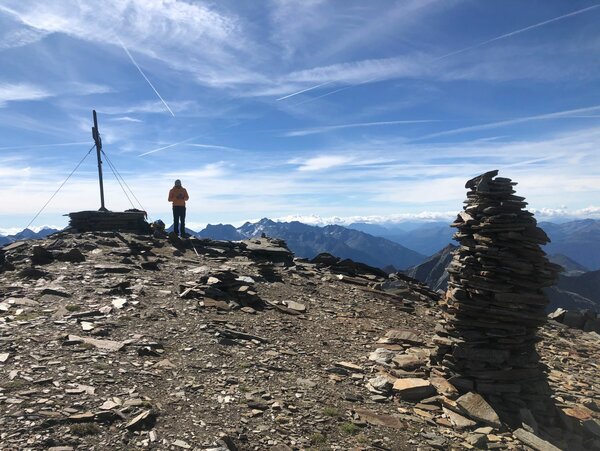Ein Steinmännchen auf einem Gipfelplateau mit einem Mensch und einem Gipfelkreuz im Hintergrund.