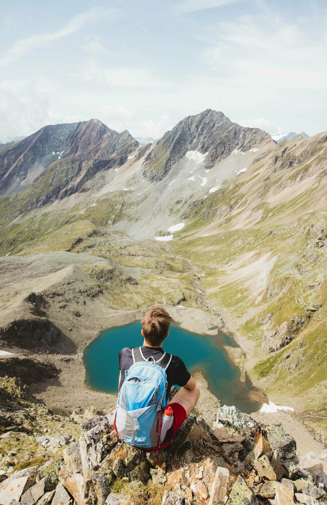 Person sitzt auf einem Stein in luftigen Höhen und blickt auf einen See hinunter.