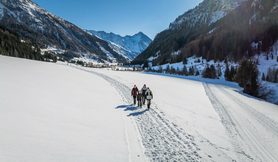 Eine Familie, bestehend aus zwei Elternteilen und zwei Kindern, wandert inmitten verschneiter Bergwelt. Die Sonne strahlt am wolkenlosen, blauen Himmel.