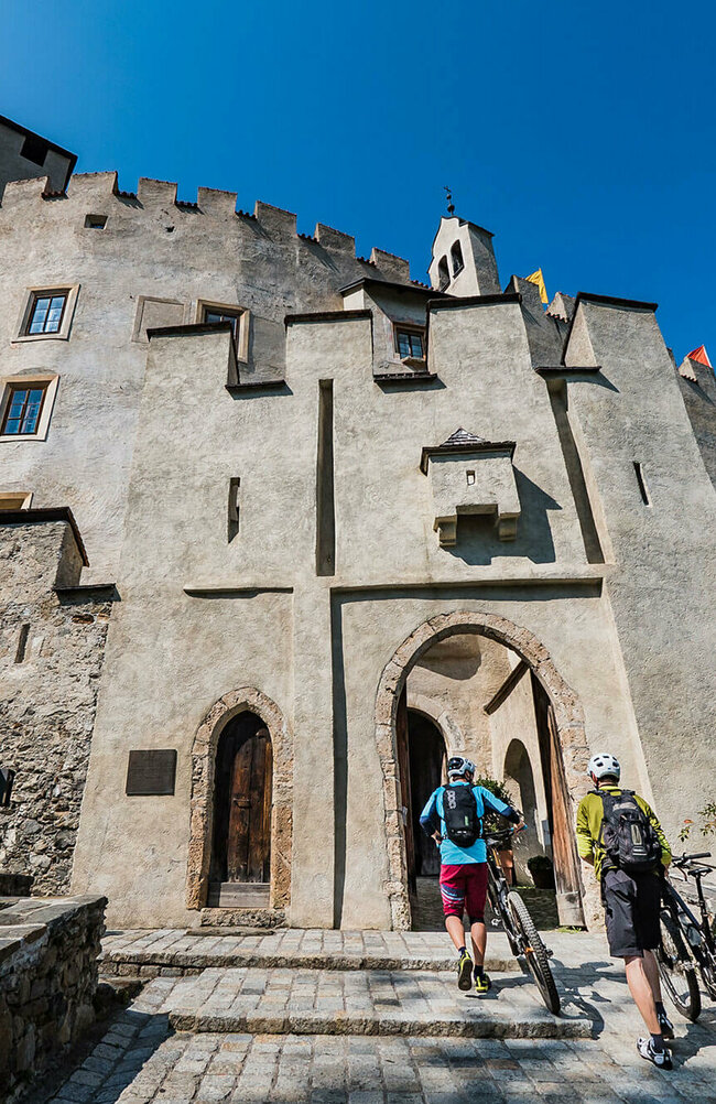 Biken Schloss Bruck Zwei Mountainbiker, die ihre Mountainbikes über einen breiten, gepflasterten Zugang zu einem alten, Schlosseingang über einen niederen Treppenabsatz schieben. Der linke Biker trägt eine weinrote, knielange Hose und eine hellblaue Radjacke, der zweite Radfahrer eine graue, knielange Hose und eine moosgrüne Jacke. Beide tragen auch einen weißen Radhelm sowie dunkle Rucksäcke. Die Schlossmauern sind vom Sonnenlicht angestrahlt und darüber blitzt blauer Himmel.