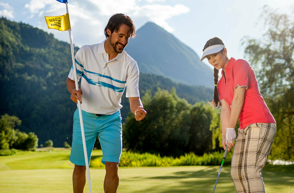 Dolomitengolf Resort Golfplatzlavantpanorama Eine Frau mit Golfschläger locht den Golfball ein im Dolomitengolf Resort. Ein Mann steht nebendran, hält die Fahne und feuert sie an.