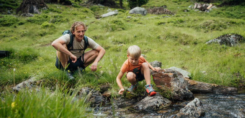 Ein Vater und ein Sohn sitzen an einem Gebirgsbach und lassen ein aus Naturmaterialien selbstgebautes Schiff im Wasser entlang gleiten.