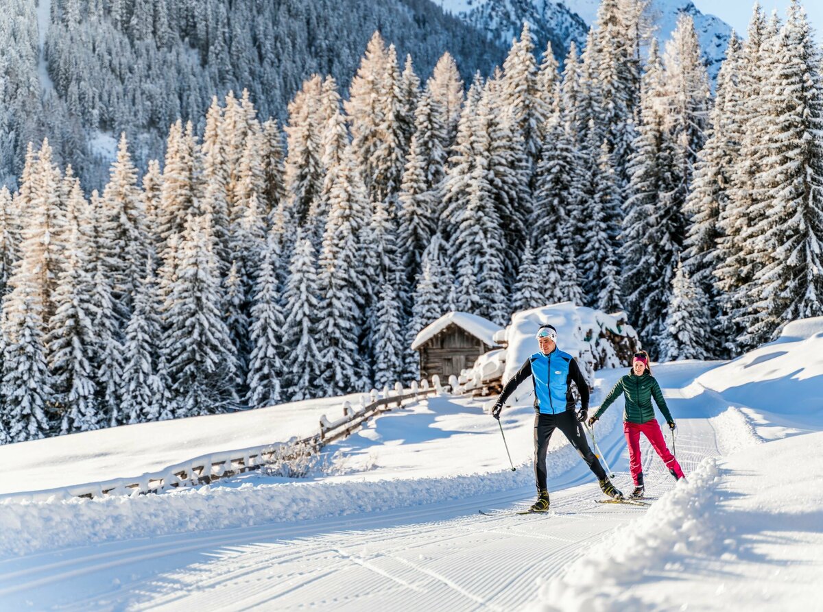 Zwei Langläufern:innen auf der Langlaufloipe in Obertilliach mit einem frisch verschneitem Wald im Hintergrund