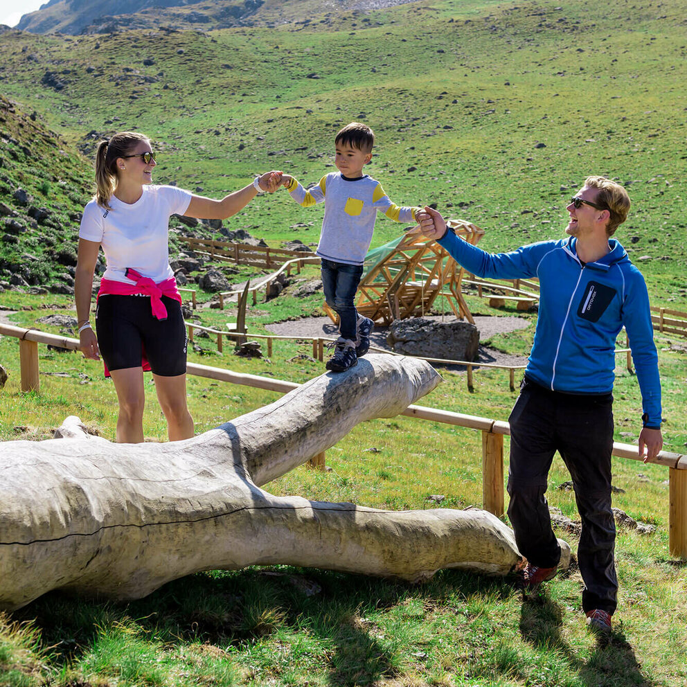 Wassermythos Ochsenlacke St. Jakob Brunnalm Eine Familie genießt die Zeit beim Wassermythos Ochsenlacke im Skizentrum St. Jakob i. D.. Ein Kind balanciert auf einem Spielgerät während er links und rechts von seinen Eltern dabei geführt wird. Die Sonne taucht die Umgebung in ein warmes Licht.
