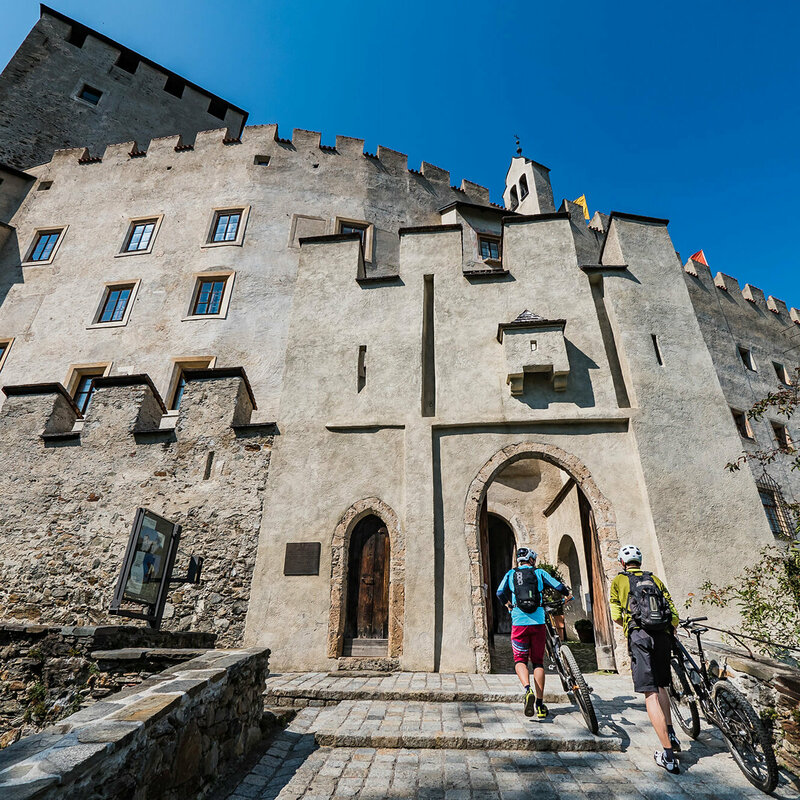 Schloss Bruck Zwei Radfahrer schieben ihre Fahrräder durch einen steinernen Eingangsbogen in das Schloss Bruck in Lienz.