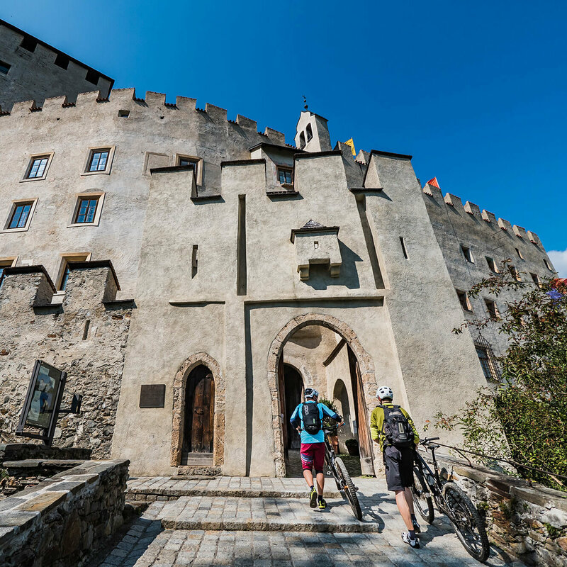Schloss Bruck Zwei Radfahrer schieben ihre Fahrräder durch einen steinernen Eingangsbogen in das Schloss Bruck in Lienz.