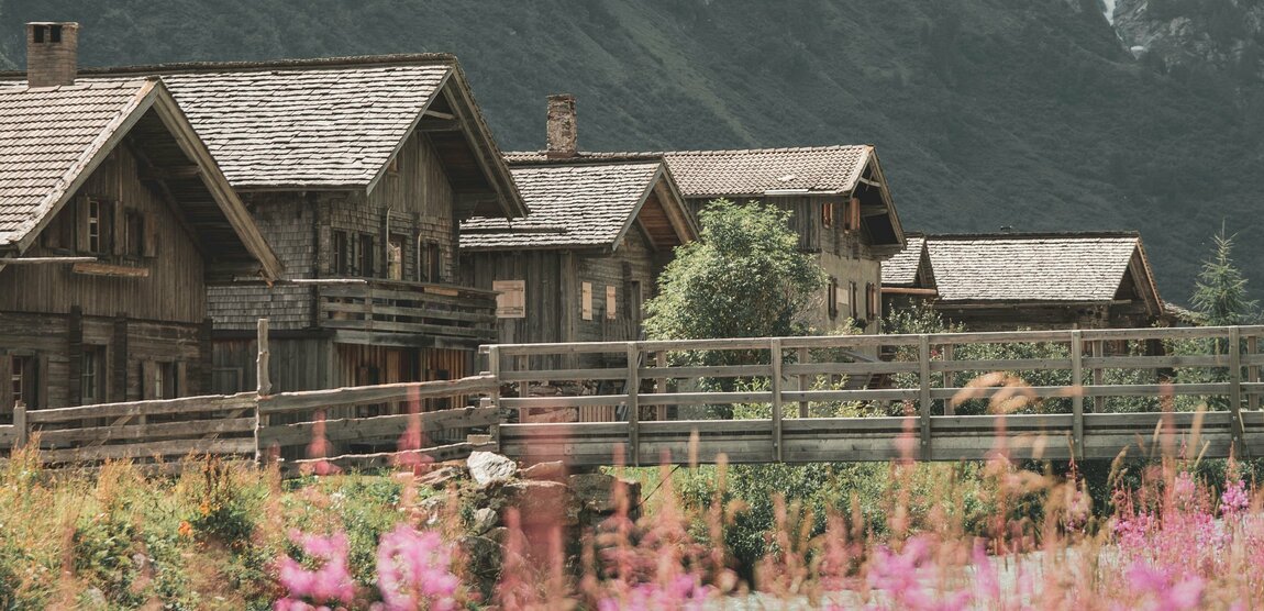 Mehrere alte Holzgebäude vor hohen, schneebedeckten Berggipfeln. Im Vordergrund wachsen rosafarbene Blumen neben einem Fluss.