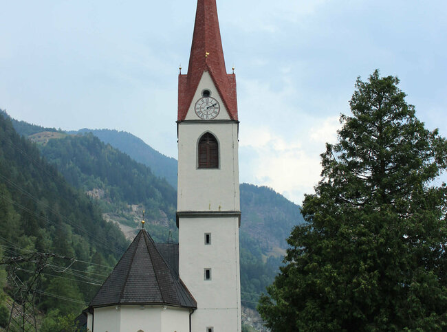 St.Johann Der rote Kirchturm der Pfarrkirche von St. Johann ragt weit in die Höhe. Daneben steht ein großer Baum und davor ein Holzzaun.