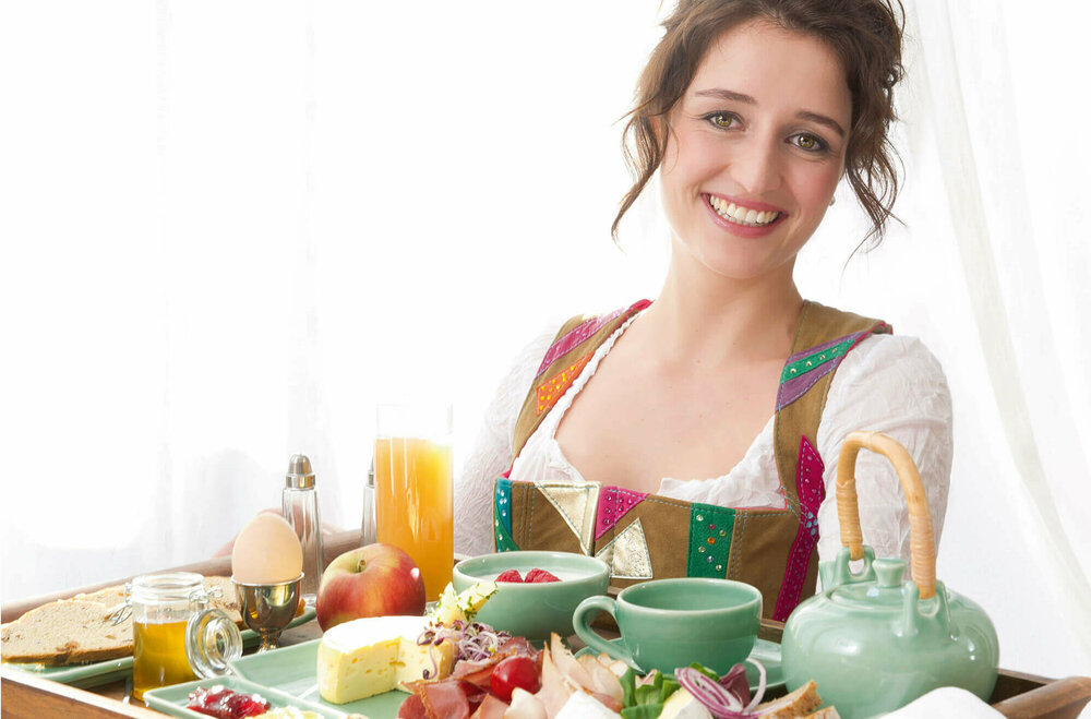 Frühstück Gutschein Dame in Dirndl serviert auf einem großen Tableau ein reichhaltiges Osttirol Frühstück mit Speck, Schinken, Käse, Ei, verschiedene Marmeladen und Honig und noch vieles Regionales mehr.