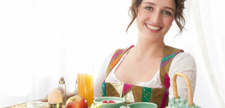 Frühstück Gutschein Dame in Dirndl serviert auf einem großen Tableau ein reichhaltiges Osttirol Frühstück mit Speck, Schinken, Käse, Ei, verschiedene Marmeladen und Honig und noch vieles Regionales mehr.