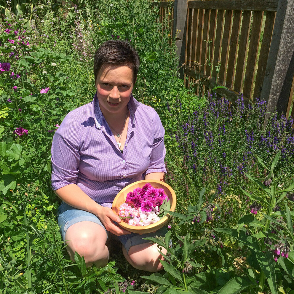 Viktoria Trager Eine Frau (Viktoria Trager) kniet in einer Blumenwiese und hält einen Holzbehälter mit Blumen gefüllt in der Hand.
