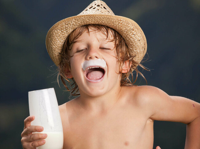 Ein Junge mit Strohhut und einem Glas Milch in der Hand. Er lacht und hat über den Lippen einen weißen Rand von der Milch.