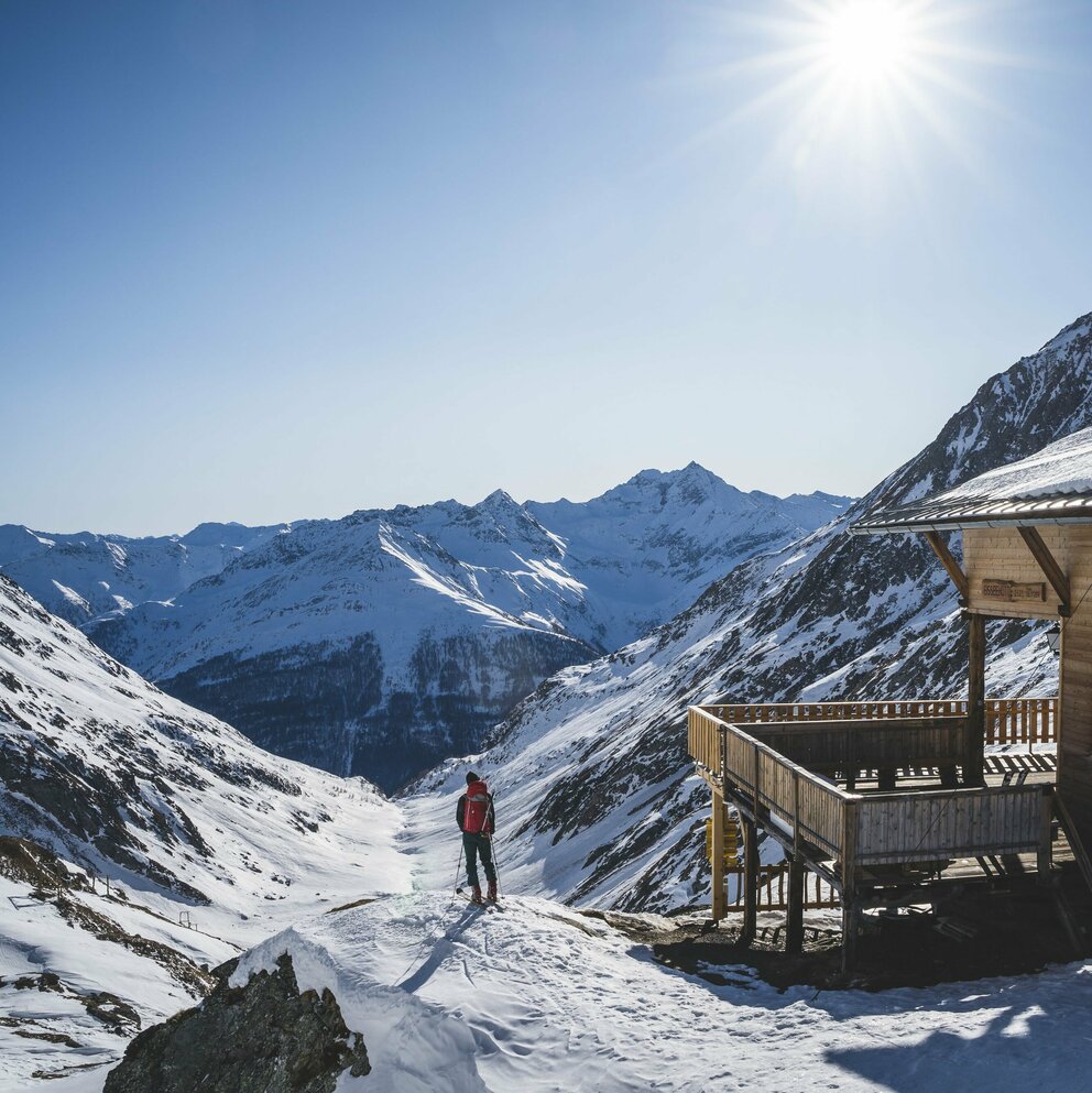 Skitour Eisseehütte Ein Skitourengeher steht neben der hölzernen Eisseehütte mit Blick talauswärts. Sie Sonne strahlt am blauen Himmel.
