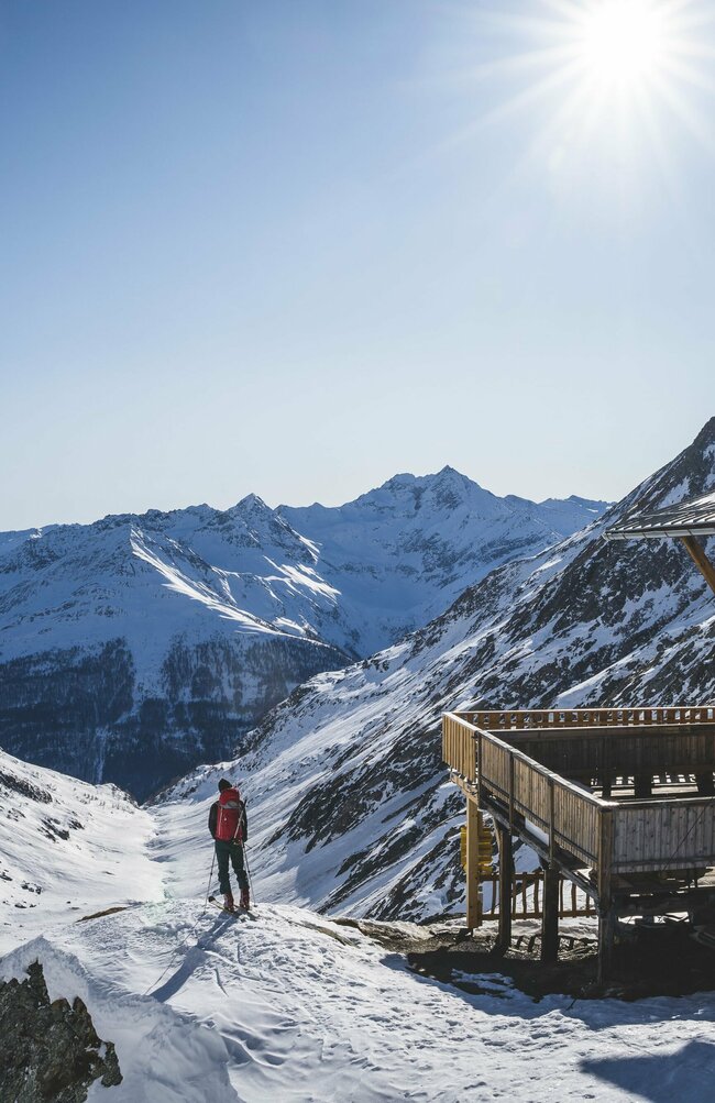 Skitour Eisseehütte Ein Skitourengeher steht neben der hölzernen Eisseehütte mit Blick talauswärts. Sie Sonne strahlt am blauen Himmel.