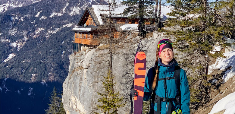 Eine Skitourengeherin steht im Schnee und hält ihre Ski in den Händen, mit der Dolomitenhütte im Hintergrund.