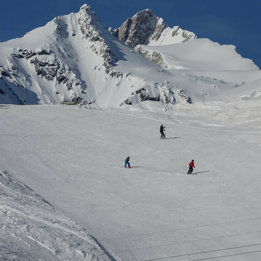 Skigebiet Bichllift Prägraten Nahaufnahme Piste Mehrere Skifahrer auf einer breiten Piste im Skigebiet Bichllift in Prägraten. Im Hintergrund ragen zwei schneebedeckte Gipfel in den blauen Himmel.