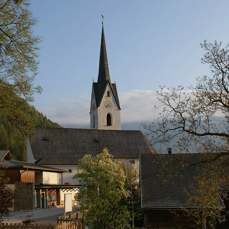 Leisach Die Kirche in Leisach inmitten des Ortskerns mit herbstlichen Bäumen und Hochnebel über dem Zettersfeld im Hintergrund.