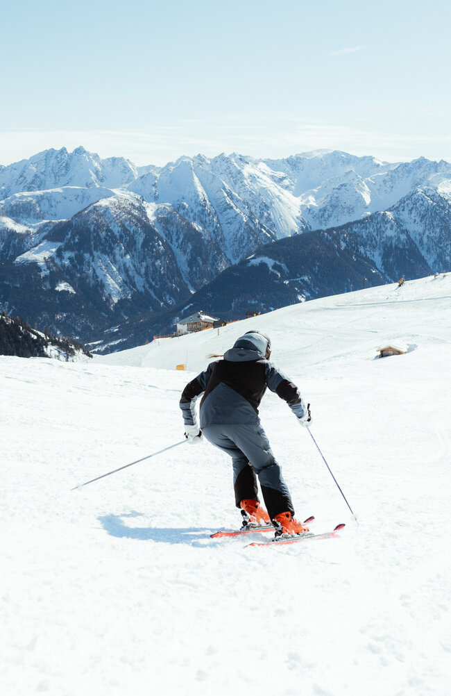 Zwei Skifahrerinnen auf einer flachen Piste oberhalb der Baumgrenze. Im Hintergrund der Taleinschnitt und ein verschneiter Bergkamm, der den Abschluss zur milchig wirkenden Bewölkung an einem Schönwettertag im Winter zeigt.