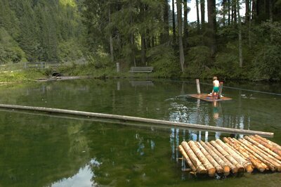 Wasserspielplatz Auentrümpfl Der Blick auf den Wasserspielplatz Auentrümpfl, in Hopfgarten i. D.. Kinder spielen auf einem Floss am See, welches durch ein gespanntes Seil von einem zum anderen Ufer gezogen werden kann.