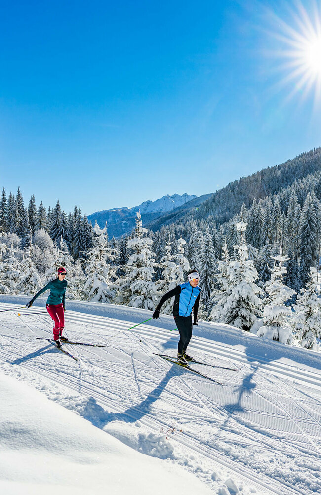 Zwei Langläufer:innen laufen bei strahlendem Sonnenschein auf der Loipe in Obertilliach mit einem frisch verschneiten Wald im Hintergrund