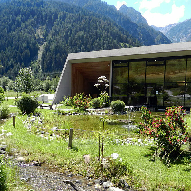 Vitalpinum Ein sanft plätschernder Bach schlängelt sich durch den Wohlfühlgarten Vitalpinum, während im Hintergrund der einladende Eingangsbereich mit dem Shop sichtbar ist.