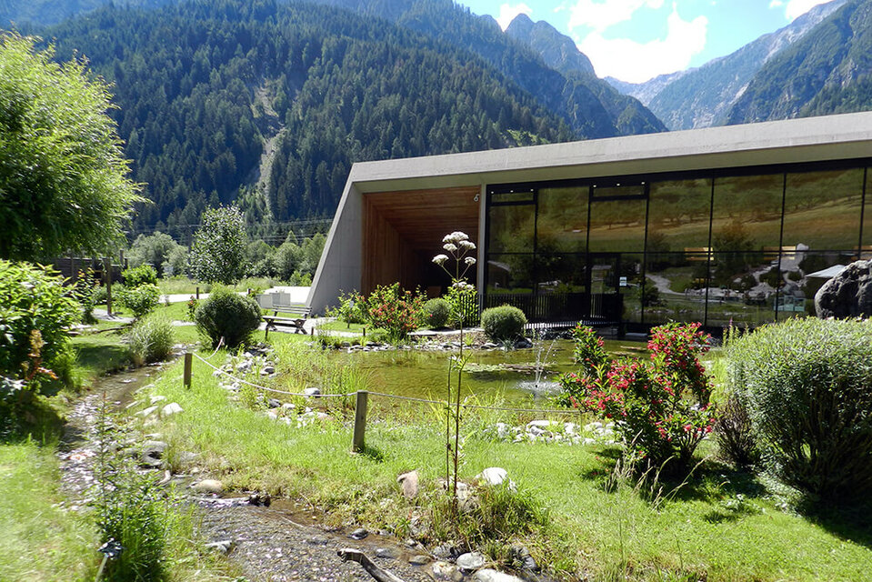 Vitalpinum Ein sanft plätschernder Bach schlängelt sich durch den Wohlfühlgarten Vitalpinum, während im Hintergrund der einladende Eingangsbereich mit dem Shop sichtbar ist.