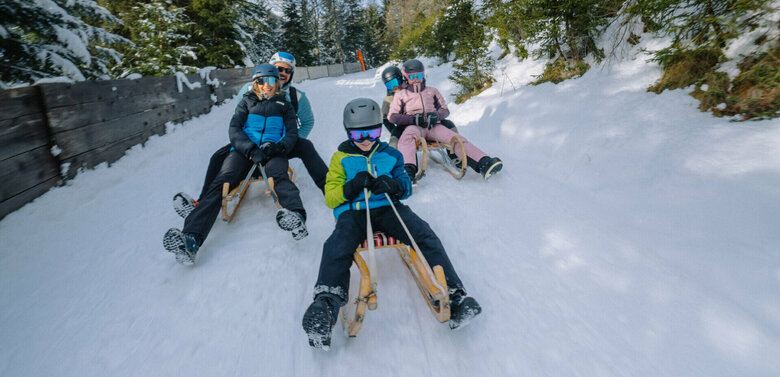 Fünf Personen in Skianzügen rodeln auf ihren Schlitten talwärts auf der Rodelbahn im Winkeltal in Außervillgraten, Osttirol.