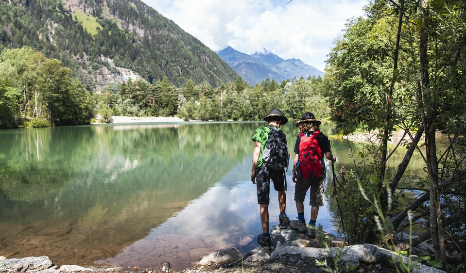 Zwei Personen in Wanderkleidung mit Rucksäcken und Hüten wandern entlang des Iseltrails an einem See in St. Johann im Walde.