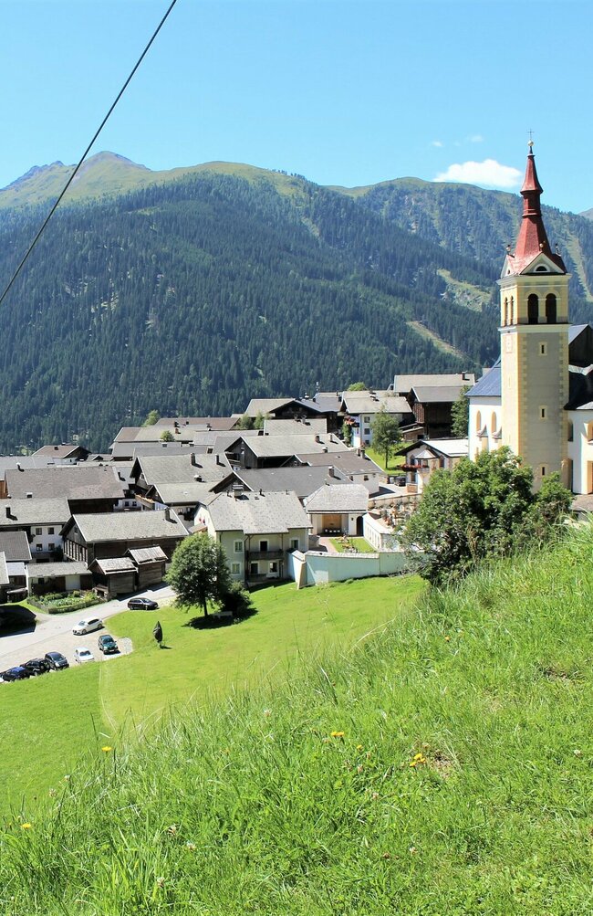 Das Dorf Obertilliach im Tiroler Gailtal, rechts im Bild die markante Kirche, links die bergwärts fahrende Gondelbahn vor alpiner Kulisse