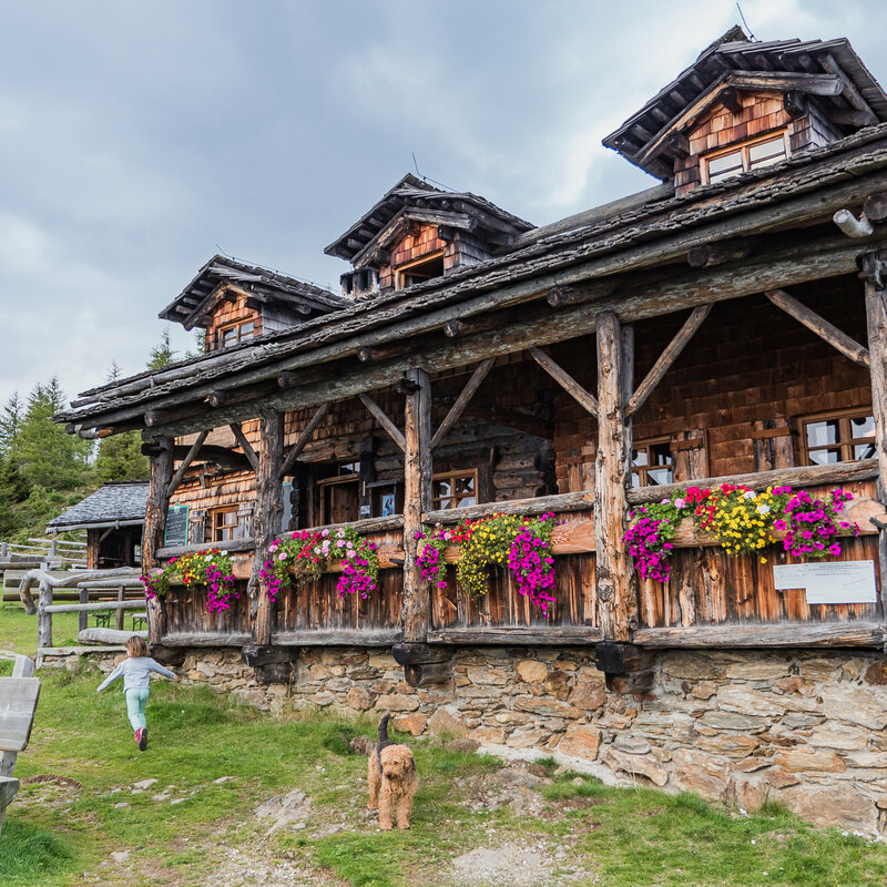 Anna Schutzhaus Das urige Anna Schutzhaus aus Holz mit Blumendeko und spielenden Kindern und einem Hund davor.