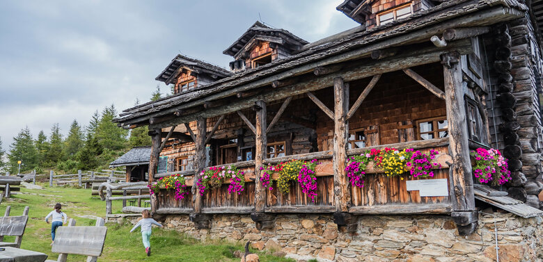 Anna Schutzhaus Das urige Anna Schutzhaus aus Holz mit Blumendeko und spielenden Kindern und einem Hund davor.