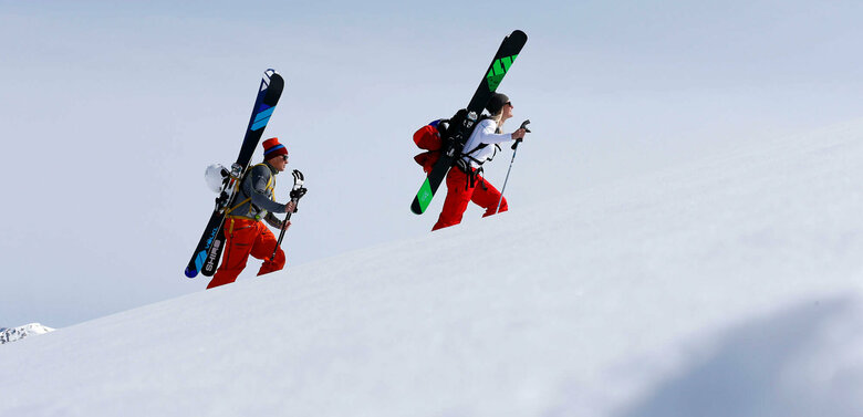 Freeriden Ein Pärchen steigt mit Ski am Rücken einen schneebedeckten Berg hinauf. Beide tragen Mützen und rote Hosen. Die Ski sind schwarz-grün und schwarz-blau und sind an einem Rucksack befestigt.