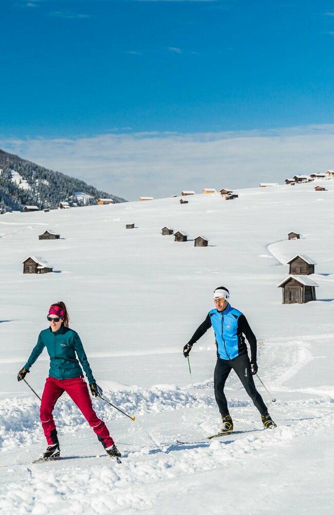 Zwei Wintersportler laufen auf ihren Langlaufskiern auf der Loipe in den Obertilliacher Feldern mit den zahlreichen kleinen Hütten und dem Dorf im Hintergrund