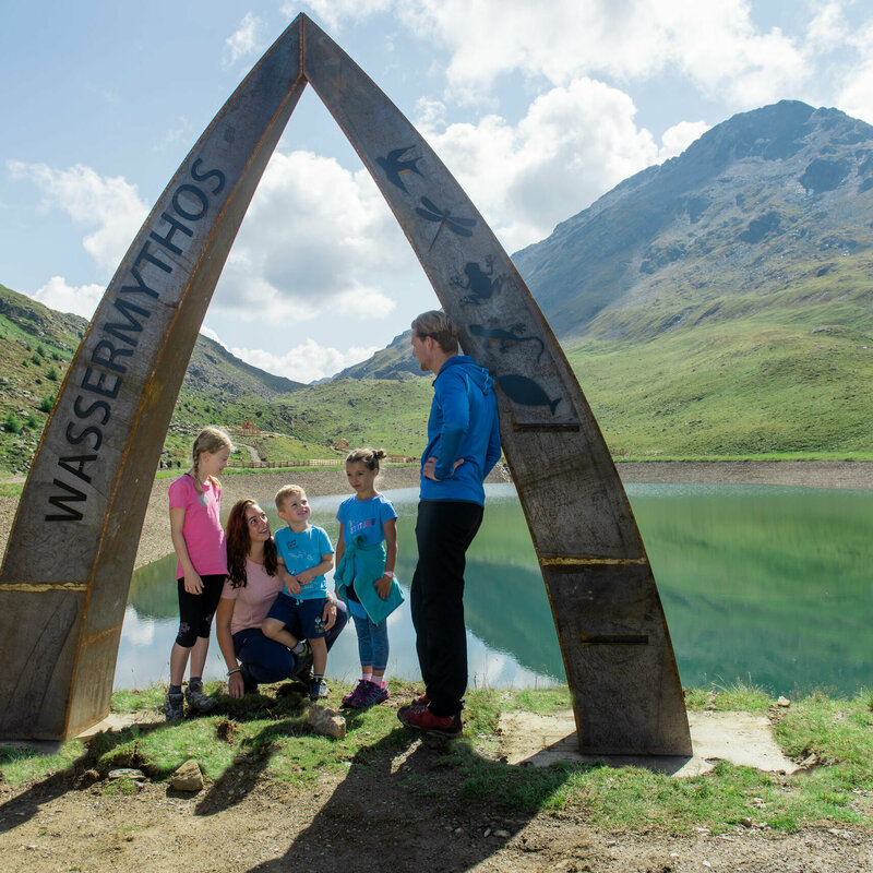 Wassermythos Ochsenlacke Eine Familie beim Wassermythos Ochsenlacke im Skizentrum St. Jakob i. D.. Sie stehen an dem kunstvollen Bogen welcher am Rande des Ufers errichtet wurde. Die Sonne lässt die umliegende Bergwelt und das Wasser in einem warmen Licht erstrahlen.