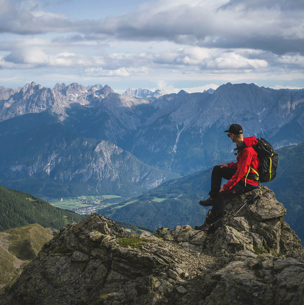 Wandern Schobergruppe Ein Wanderer mit roter Jacke, schwarzem Rucksack und Kappe sitzt auf einem Felsbrocken in der Schobergruppe und blickt ins Tal Richtung Oberlienz.