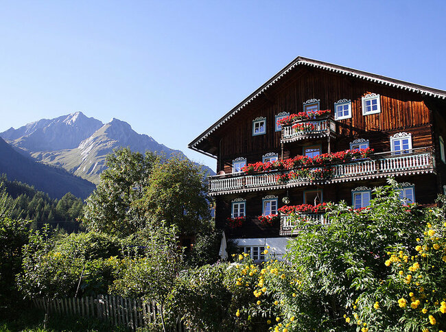altes Bauernhaus in Kals/Burg Blick auf ein altes Bauernhaus in Kals mit roten Blumen auf den Balkonen und einem Berg im Hintergrund.
