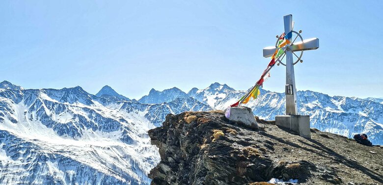 Figerhorn Frühling Gipfelkreuz am Figerhorn im Frühling mit noch winterlichen Berggipfeln im Hintergrund.